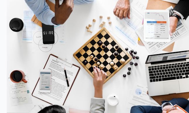 Top view of business colleagues around a table doing work. In the center of the table is a chess board that one person is playing.