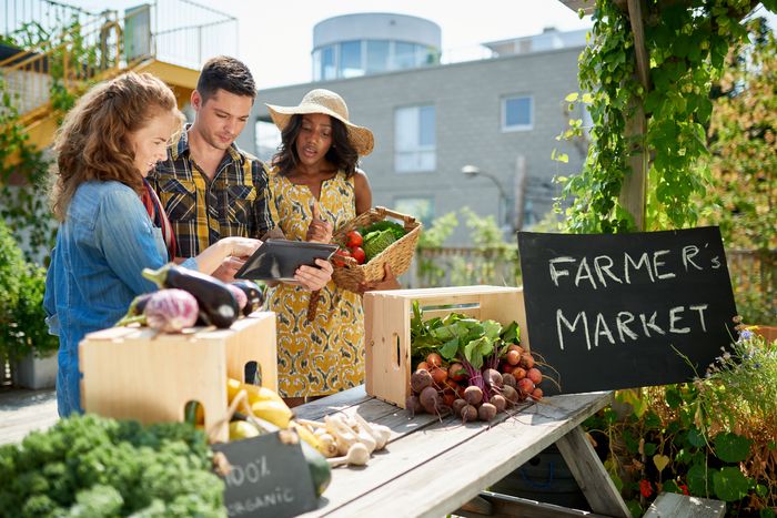 A man holding a tablet with two women looking at the tablet discussing circulat economy for food while at a farmers market. They are surrounded by fruit and vegetables.
